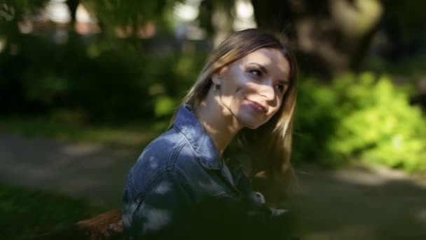 若い美しい女性は公園のベンチでリラックスして彼女の髪をまっすぐにします 木の緑の葉を通して撮影 — ストック動画