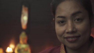 Kırmızı sabahlıklı Taylandlı masöz gülümsüyor. Mumlar yanar ve arkada bir Buda heykeli durur. Yakın plan..