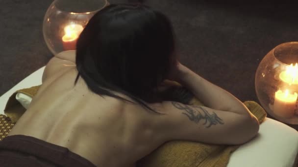 美丽的女孩躺在床上做按摩 床边的蜡烛在燃烧 美丽的背影和手臂上的纹身 后续行动 — 图库视频影像