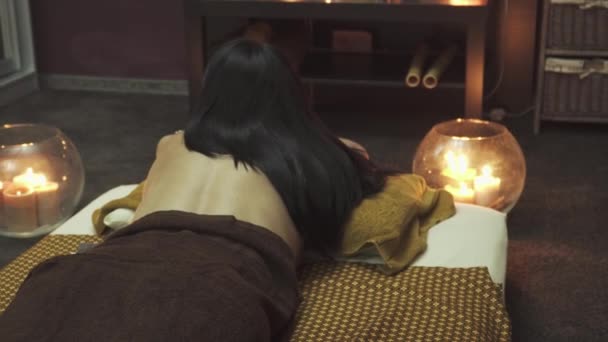 漂亮的黑头发躺在按摩床上 床边的蜡烛在燃烧 相机从上至下移动 — 图库视频影像