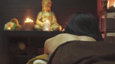 Masaj salonundaki güzel kız elinde mumlarla bir masanın önünde. Masanın üzerinde bir Buda heykeli var, orkideler yalan söylüyor ve mumlar yanıyor. Kamera sağdan sola hareket eder.
