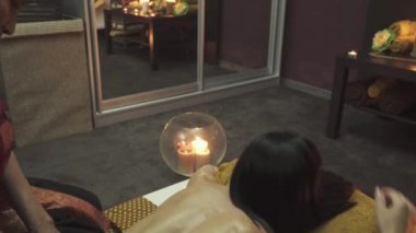 Thai Spa 'daki masöz kızın üstüne oturur ve sırt masajı yapar. Masaj yatağının yanındaki köşede mumlar yanıyor. Kamera yukarıdan aşağıya hareket eder.