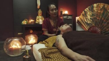 Taylandlı masöz güzel bir kıza kafa masajı yapıyor. Mumlar yanar ve arka planda bir Buda heykeli durur..