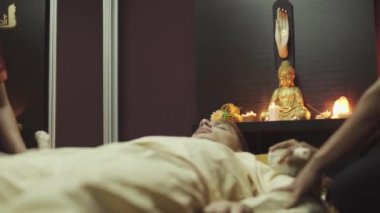 Adam iki Taylandlı kadından masaj alıyor. Masaj salonunda. Çocuk masaj yatağında yatıyor. Mumlar masada yanıyor ve arka planda Buda heykeli var..