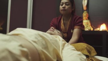 Güzel Taylandlı masöz masaj salonundaki bir adamın boynuna yavaşça masaj yapıyor. Buda heykeli arka planda mumlarla bir masanın üzerinde duruyor.