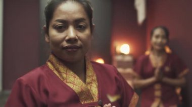 Masaj salonunda kırmızı sabahlık giymiş iki güzel Taylandlı masöz. Masanın arka planında mum yanıkları var. Kapat..