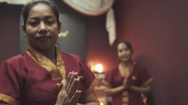 Masaj salonunda kırmızı sabahlık giymiş iki güzel Taylandlı masöz. Masanın arka planında mum yanıkları var. Kapat..