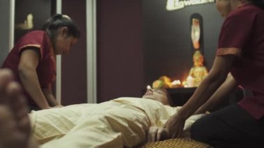 Mutlu adam masaj yatağında yatıyor ve iki Taylandlı masözden masaj alıyor. Mumlar masada yanıyor ve arka planda bir Buda heykeli var.,