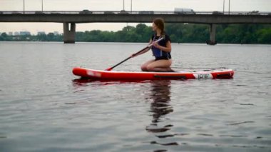 Genç ve güzel esmer bir kanoya biner ve nehir boyunca kürek çeker. Arka planda otomobil köprüsü.