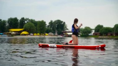 Genç güzel kız bir kanoya diz çöker ve nehir boyunca yelken açar. Arka planda şehir plajı.