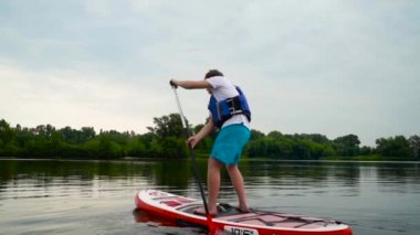 Genç adam nehir boyunca bir kanonun üzerinde ayakta yüzer. Adam küreği elden ele hareket ettirerek kanoda yüzer. Adam küreklerini başının üstüne kaldırdı..