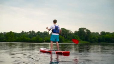 Genç adam nehir boyunca bir kanonun üzerinde ayakta yüzer. Adam küreği elden ele hareket ettirerek kanoda yüzüyor..