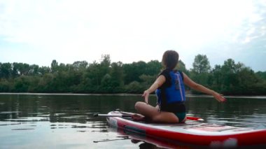 Güzel esmer bir kanoda oturuyor. Genç kız nehirde yoga yapıyor. Kız ellerini yana açıyor.