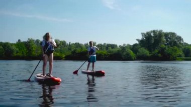 Güneş gözlüklü güzel bir sarışın ve kanoların üzerinde birlikte yüzen yakışıklı bir adam. Arka planda alçak ağaçlar var..