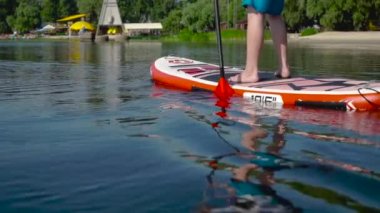 Sporcu çocuk kanoya binip yüzüyor. Sadece kanodaki bir adamın bacakları görünür. Önünde şehir plajı var..