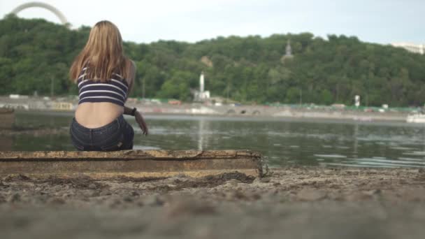 年轻的女人坐在河边 女人在户外消磨时光 女人们欣赏美丽的风景 从后面开枪 — 图库视频影像