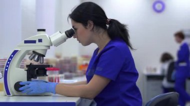 Laboratuvar teknisyeni laboratuarda mikroskoba bakıyor. Ekip arka planda çalışıyor.