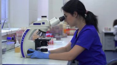 Laboratuvar teknisyeni laboratuarda mikroskoba bakıyor. Ekip arka planda çalışıyor.