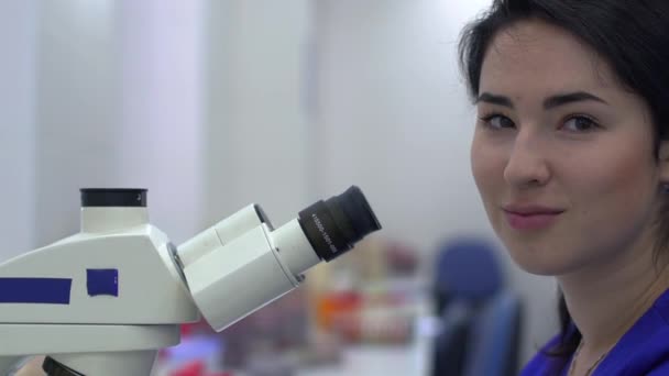 顕微鏡を使って研究室で分析をしている若い女性 女性はカメラを見て笑っている 医療化学反応だスローモーション — ストック動画