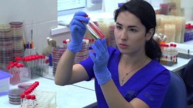 Mavi üniformalı genç bir kadın test tüpleri için raftan iki tüp alıyor ve renk ve sıvı yoğunluğunu kontrol ediyor, sonra da gülümseyen kameraya bakıyor. Laboratuvarda araştırma yapıyorum. Bilim, meslek, sağlık. Yavaş çekim