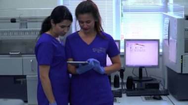 Mavi üniformalı iki kadın laboratuvar çalışanı modern laboratuarda bilgisayarların ve makinelerin önünde tabletle araştırma yapmayı tartışıyor. Bilim, meslek, sağlık. Yavaş çekim