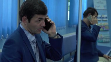 Resmi giyinen yetişkin adam modern bir özel klinikte koridorda oturmuş cep telefonuyla analiz sonuçlarını bekliyor. Hastanın aynadaki yansıması. Sağlık hizmetleri kavramı