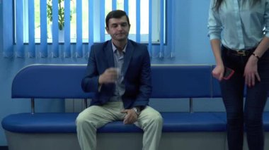 Bir kadın sandalyeye oturur ve sıranın gelmesini bekler. Mavi renkli bir oda. Adam su içiyor. Kadın cep telefonuyla oynuyor.