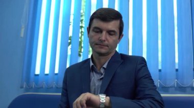 Bir bekleme odasında oturup elindeki saate bakan takım elbiseli yetişkin bir adam portresi..