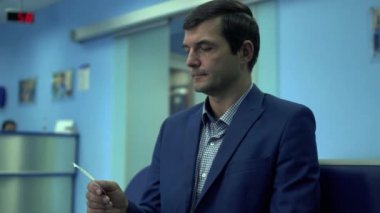 Resmi giyinen yetişkin adam modern özel kliniğin koridorunda oturmuş biletini elinde tutmasını bekliyor. İş adamı saate bakıyor.