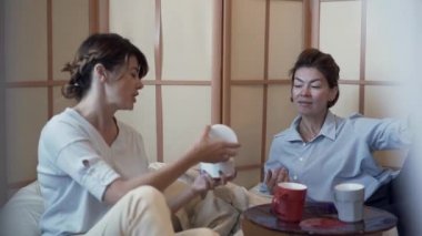 İki sevimli olgun kadın rahat yastık sandalyelerde küçük masada oturan kahve içme ve kozmetik hakkında konuşurken. Genç bayan arkadaşına yeni bir krem gösterir. Vücut bakımı. Yavaş çekim.