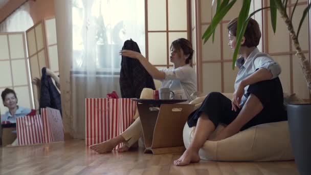 Twee volwassen vrouwen zitten aan de kleine tafel op comfortabele kussen stoelen praten en delen. Jongere dame toont haar vriend een nieuwe jas nemen van het uit de zak. Slow Motion — Stockvideo