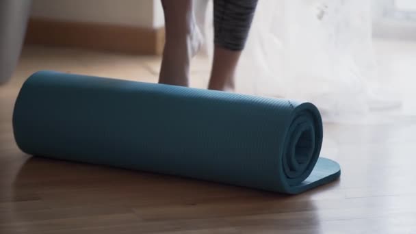 Mujer no reconocida desenrollando la esterilla de yoga acostada en el suelo usando su pie. La chica que se prepara para una clase de yoga o meditación en casa o en el estudio. Estilo de vida saludable — Vídeos de Stock