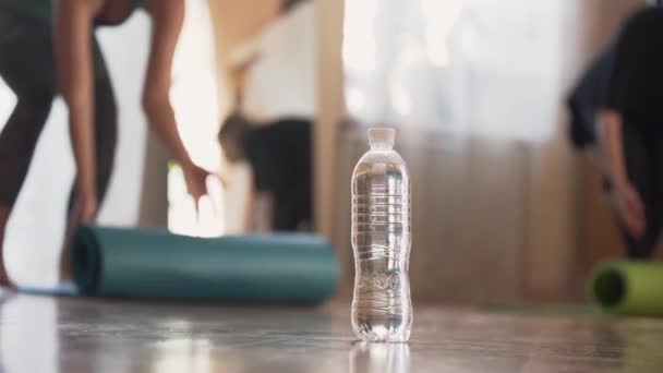 Dos mujeres desenrollando la esterilla y sentadas en ella preparándose para la clase de yoga o meditación en casa o en el estudio. La botella con el agua de pie en el suelo en primer plano. Estilo de vida saludable — Vídeos de Stock