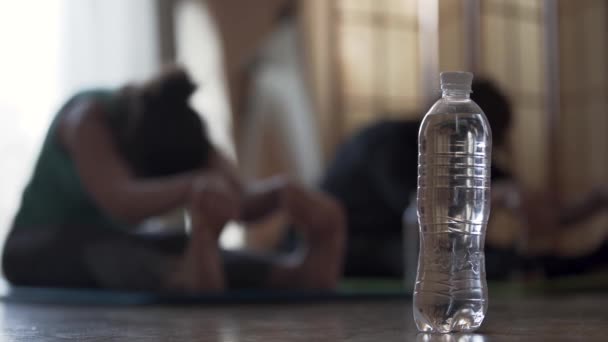Дві з фокусу дівчата роблять вправи, сидячи на підлозі. Пляшка води на передньому плані. Здоровий спосіб життя, відпочинок, збереження форми . — стокове відео
