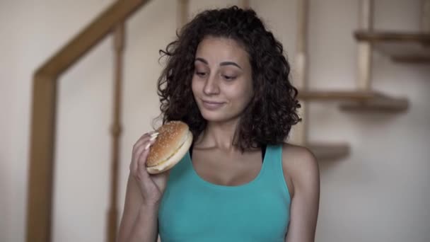 Portrait de mignonne jeune femme bouclée tenant un gros hamburger et un petit haltère dans les mains en essayant de choisir entre un mode de vie sain et une nourriture savoureuse. Choix, concept de dilemme . — Video