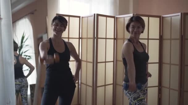 Two happy mature girlfriends doing exercises with dumbbells while looking at the camera. Hand exercise. Healthy lifestyle, recreation. — Stock Video