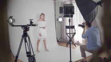 Fotoğraf çekiminin sahne arkası. Profesyonel erkek fotoğrafçı stüdyoda beyaz arka plan üzerinde sandalyede oturan sevimli zarif kadın fotoğrafları çekiyor. Moda dergisi stüdyo photoshoot.