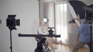 Fotoğraf çekiminin sahne arkası. Ünlü erkek fotoğrafçı stüdyoda beyaz arka plan üzerinde sandalyede oturan çekici zarif kadın fotoğraflarını çekiyor. Moda dergisi stüdyo photoshoot.