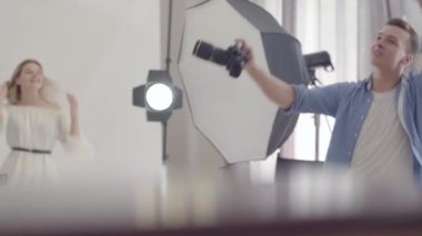 Fotoğraf çekiminin sahne arkası. Profesyonel erkek fotoğrafçı beyaz arka plan üzerinde stüdyoda fotoğraf çekmeden önce beyaz elbiseli bir model ile dans. Fotoğraf, meslek, fotoğraf çekimi.