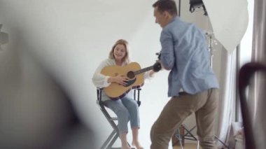 Fotoğraf çekiminin sahne arkası. Ünlü profesyonel fotoğrafçı stüdyoda beyaz arka plan üzerinde sandalyede otururken gitar çalan genç kadının fotoğraflarını çekiyor. Moda stüdyosu photoshoot.