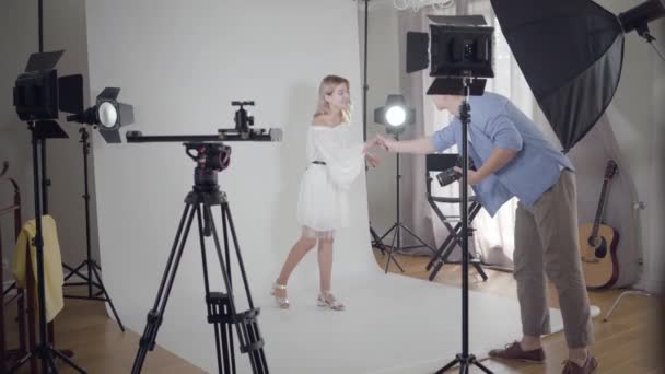V zákulisí focení. Fotograf se třese s modelkou, která stojí na bílém pozadí ve studiu před natáčet. Módní fotoalbum. — Stock video