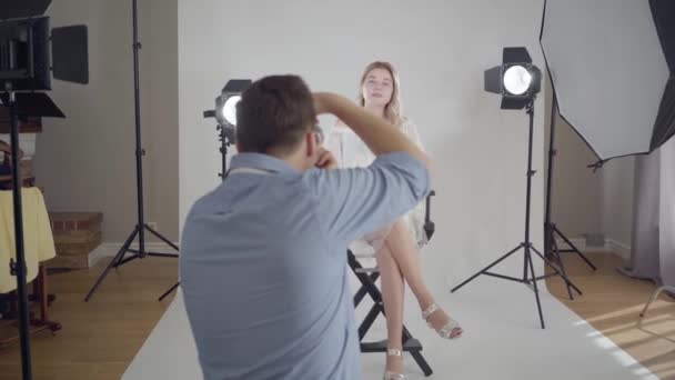 Famoso fotógrafo masculino tomando fotos de una atractiva mujer elegante sentada en la silla sobre fondo blanco en el estudio. Entre bastidores de la sesión de fotos. Sesión de fotos de estudio de revista de moda . — Vídeo de stock