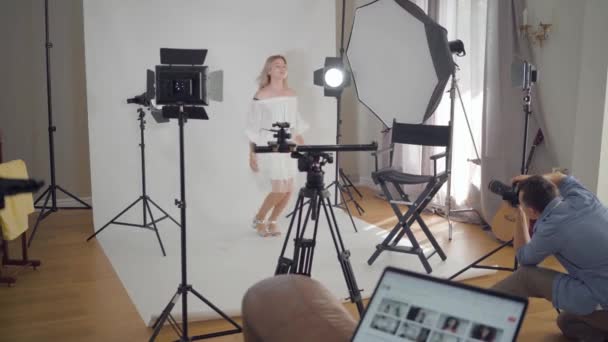 Entre bastidores de la sesión de fotos. Fotógrafo profesional tomando fotos de una joven modelo saltando sobre fondo blanco en el estudio. Sesión de fotos de estudio de revista de moda . — Vídeos de Stock