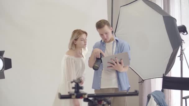 Entre bastidores de la sesión de fotos. Joven fotógrafa exitosa muestra ejemplos de su trabajo en una computadora portátil a una chica mientras está de pie sobre fondo blanco en el estudio. Sesión de fotos de estudio de revista de moda . — Vídeos de Stock