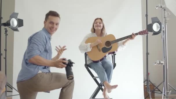 За лаштунками фотосесії. Професійний веселий фотограф фотографує молоду дівчину, яка грає на гітарі, сидячи на стільці на білому тлі в студії. Фотосесія студії моди . — стокове відео