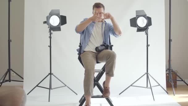 Profesionální mužský fotograf sedí na židli a zaměřuje se jeho prsty na kameru ve studiu na bílém pozadí. Fotografie, profese, Foto sezení. Pohled zepředu — Stock video