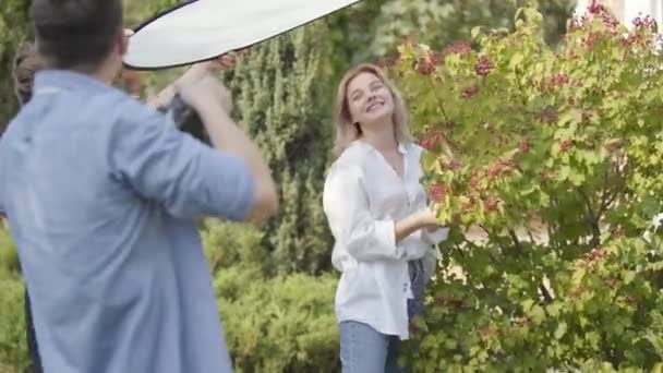 Młoda pozytywna dziewczyna w białej koszulce i dżinsy stwarzające w pobliżu krzewu jagodowego dla młodego mężczyzny z kamerą na zewnątrz. Mężczyzna fotograf robienia zdjęć atrakcyjnej kobiety w parku — Wideo stockowe