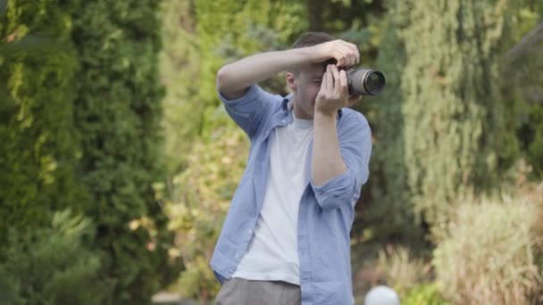 Retrato de un joven fotógrafo varón confiado tomando fotos con la cámara de pie al aire libre. Fotografía, profesión, sesión de fotos . — Vídeo de stock