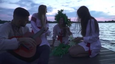 Üç kız ve gitarlı bir adam iskelede oturuyorlar. Beyaz ulusal kostümlü kızlar. Bir kızın başında yabani çiçek çelengi. Ulusal tişörtlü adam gitar çalıyor..
