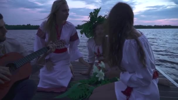 三个女孩和一个拿着吉他的男人坐在码头上 穿着白色民族服装的女孩 一个女孩头上戴着野花的花环 穿着国产服的家伙弹吉他 — 图库视频影像